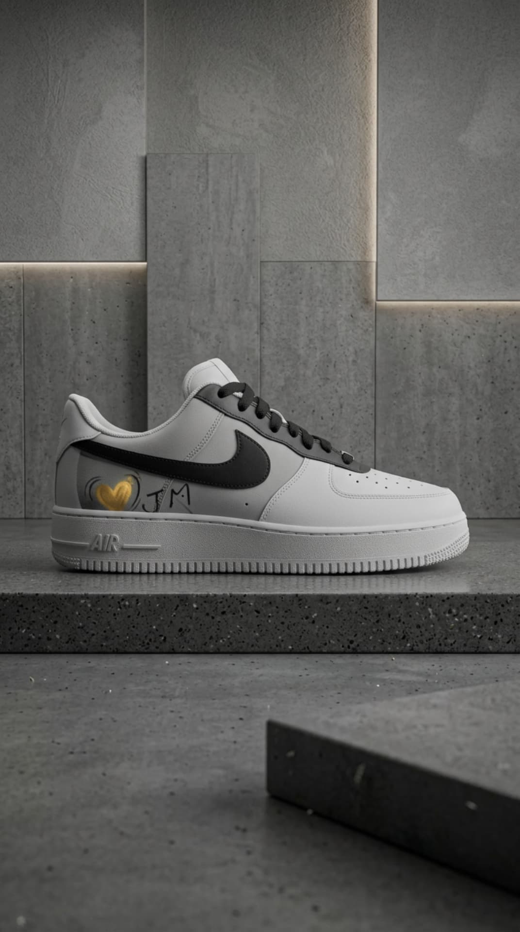 Grey and white Air Force 1 with a gold heart and JM script on a concrete ledge in a muted grey room