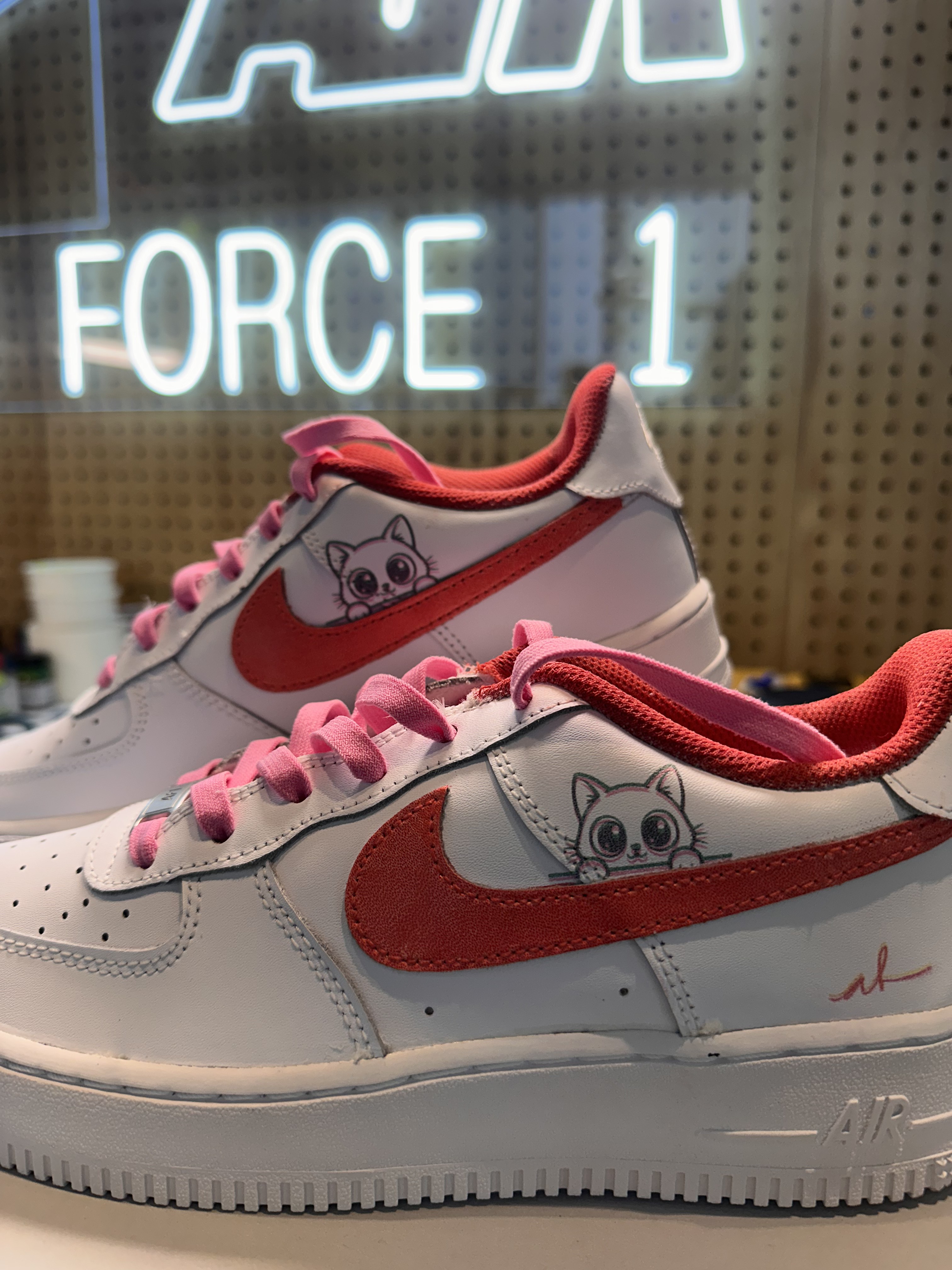Custom white Air Force 1s with pink laces and hand-drawn cat artwork under an Air Force 1 neon sign