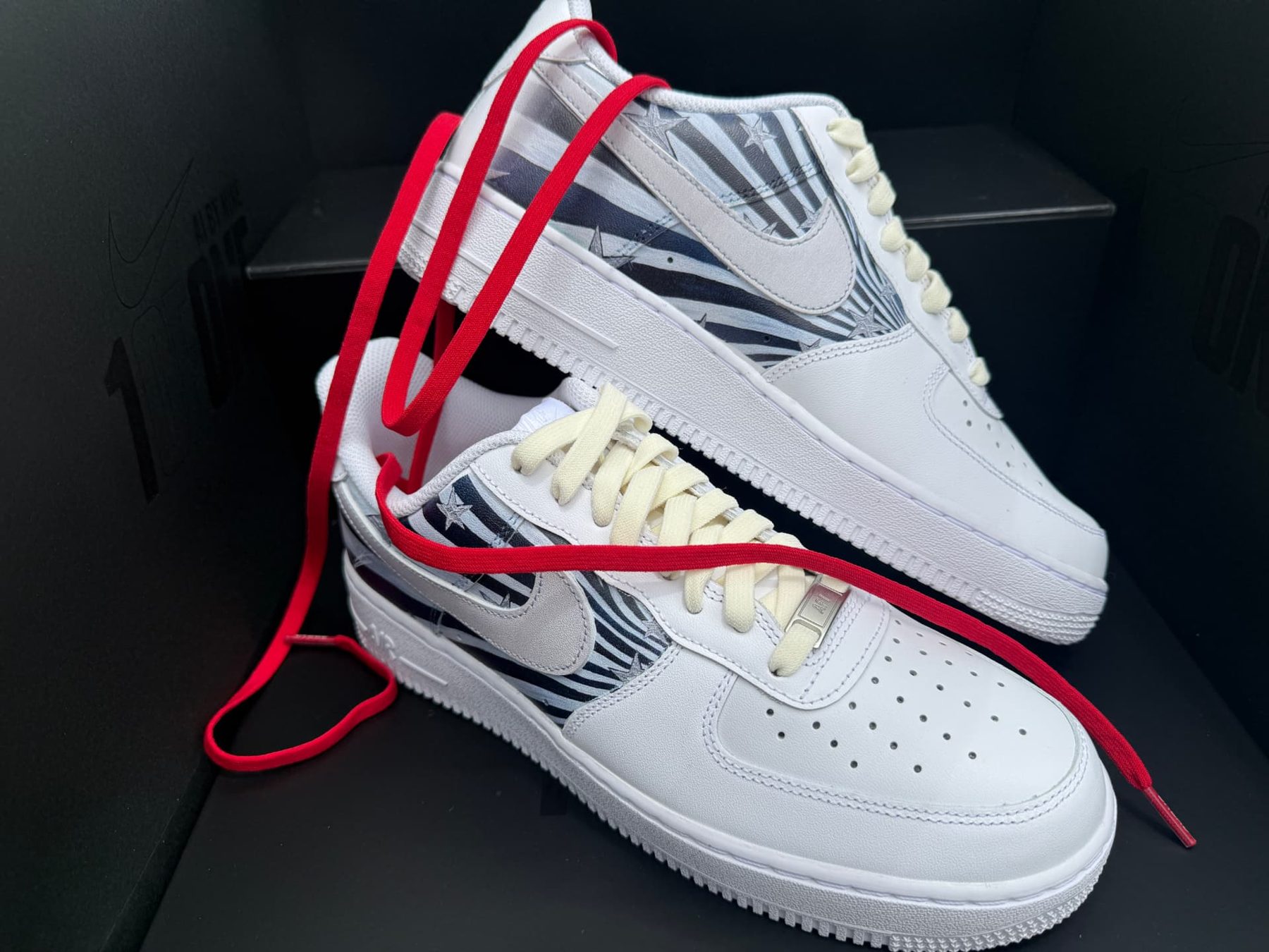White Air Force 1s with stars and stripes print panels and loose red laces on a black 1ofONE display