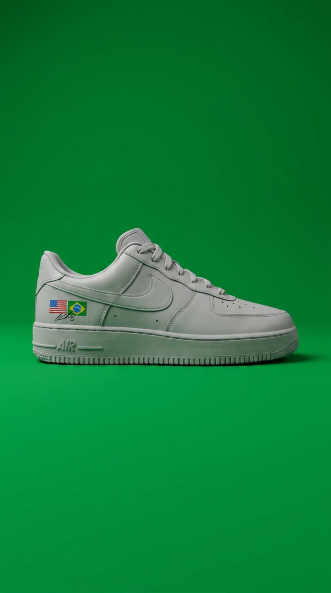 All-white Air Force 1 with USA and Brazil flag decals and a signature, against a vivid green backdrop