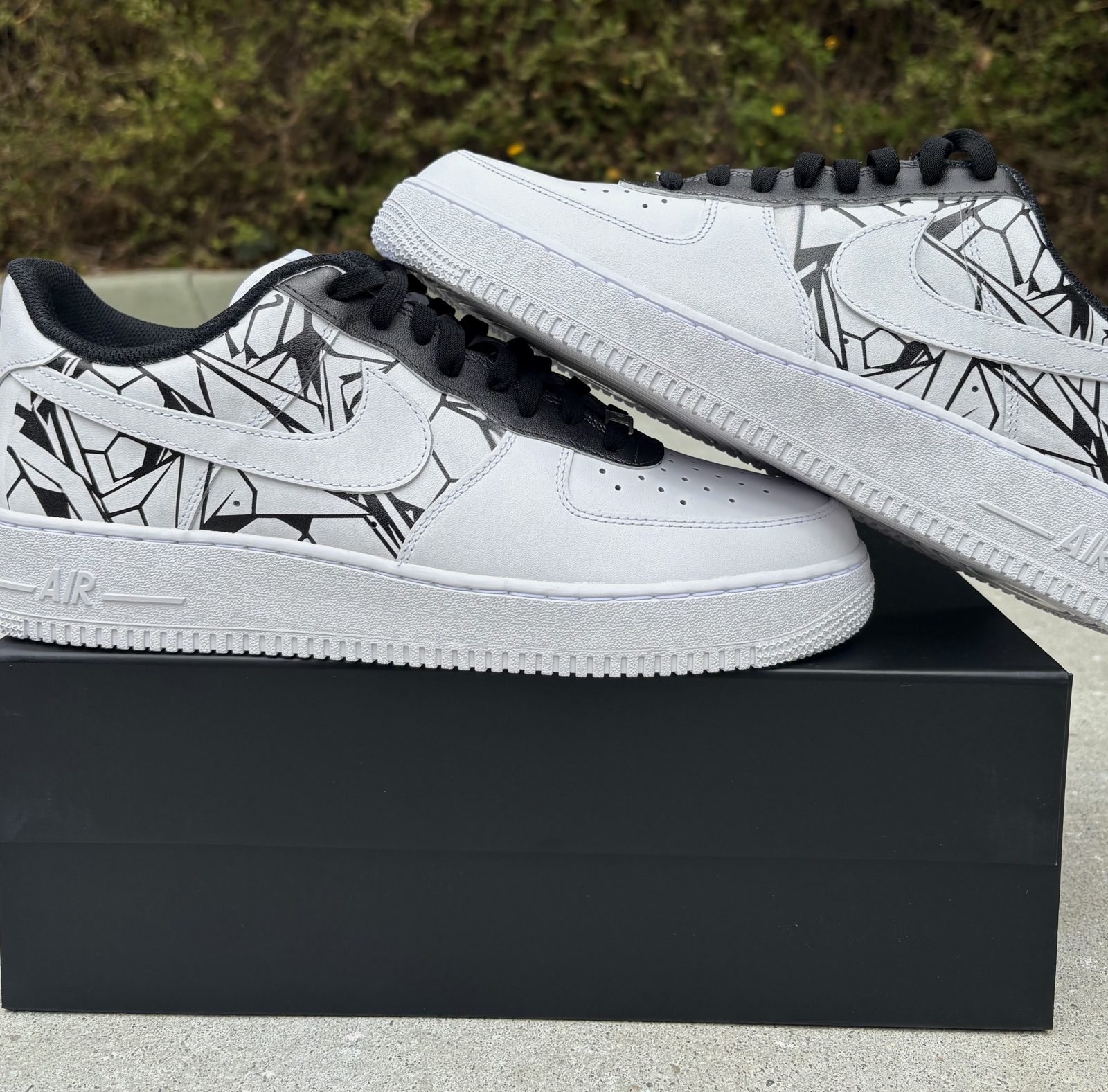 Pair of white Air Force 1s with black geometric line artwork and black laces resting on a 1ofONE box
