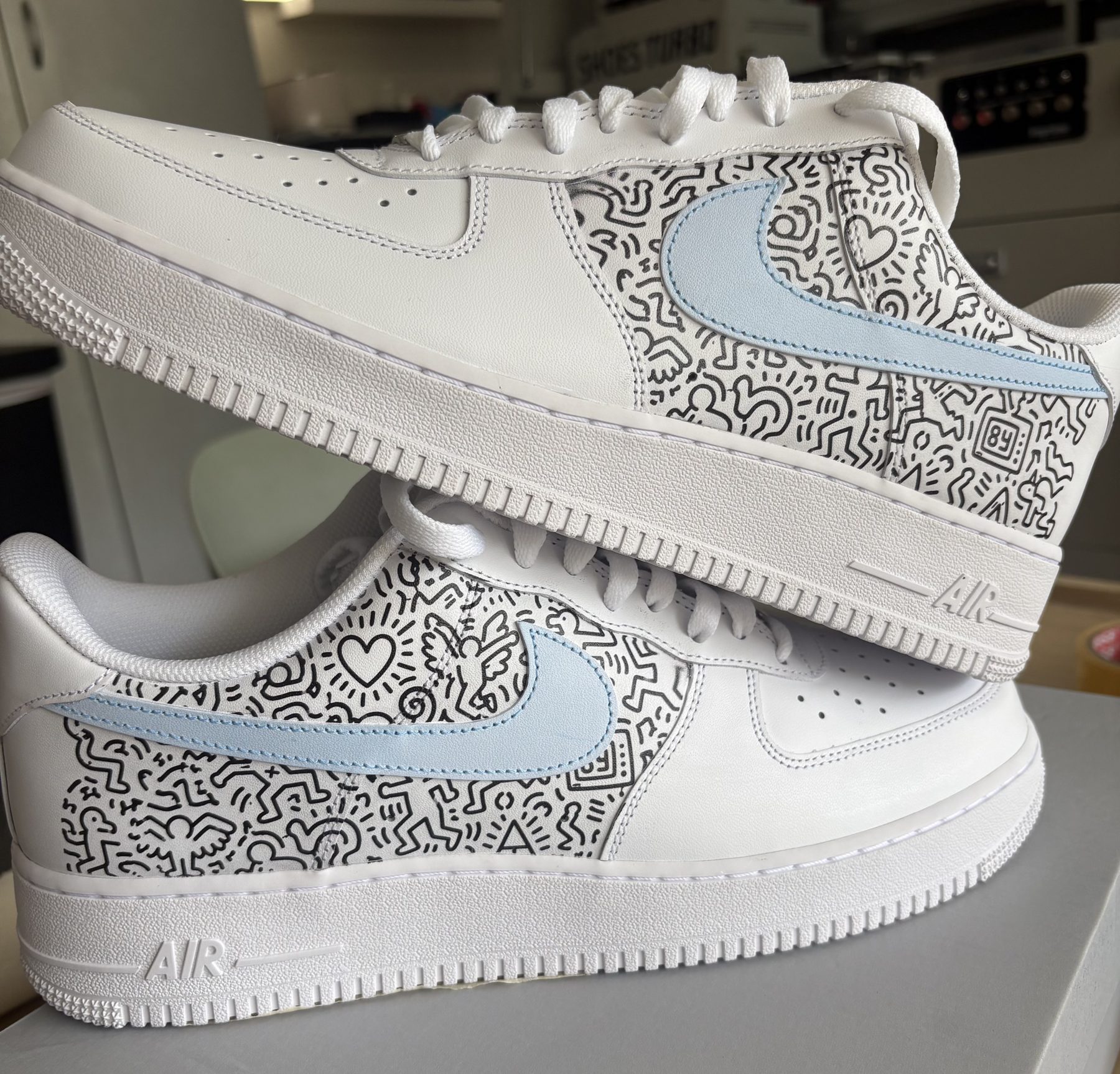 Pair of white Air Force 1s with black doodle line art and pale blue swooshes