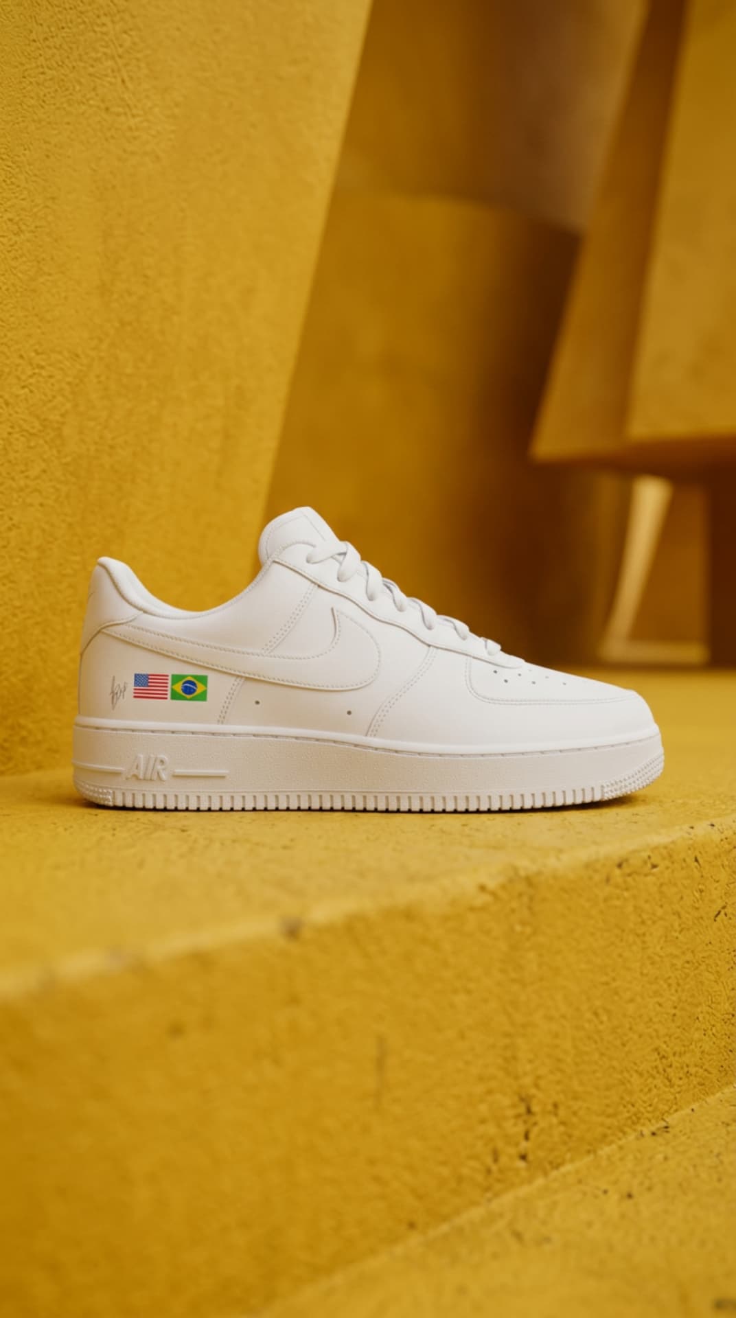 All-white Air Force 1 with USA and Brazil flag decals on a yellow concrete step in warm sunlight