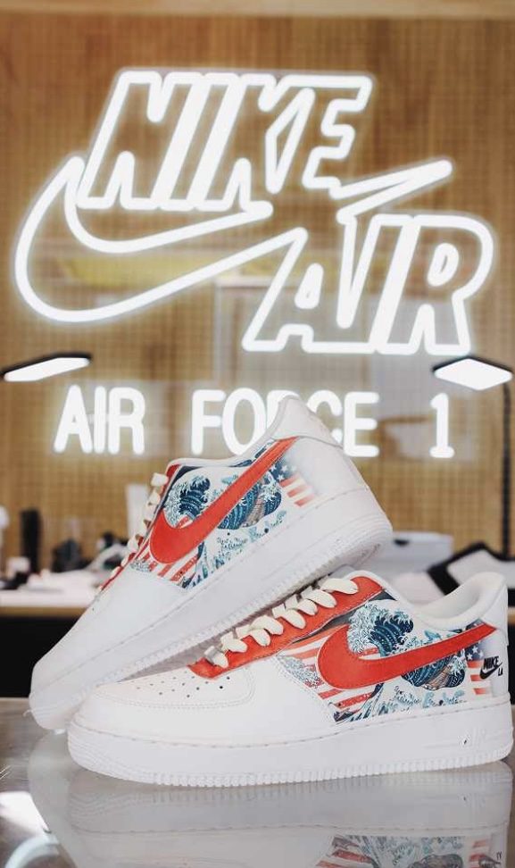 Pair of white Air Force 1s with a great wave and stars-and-stripes print under a Nike Air Force 1 neon sign