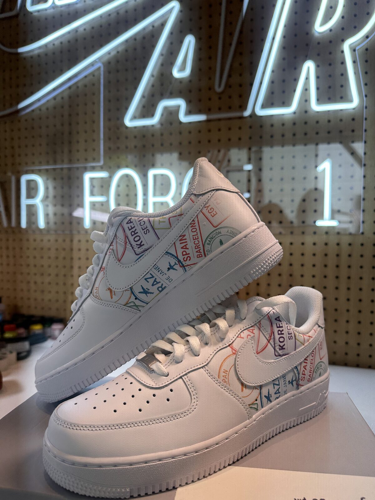 Pair of white Air Force 1s with multicolored passport stamp print displayed under a Nike Air Force 1 neon sign