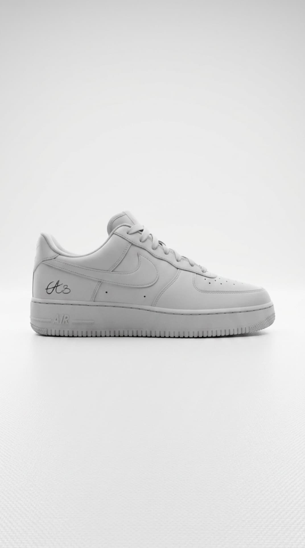 All-white Air Force 1 with a small script signature on the heel panel against a clean white studio backdrop
