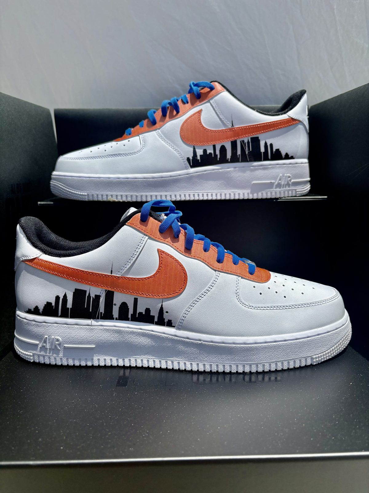 Pair of white Air Force 1s with an orange suede swoosh, blue laces and a black city skyline print