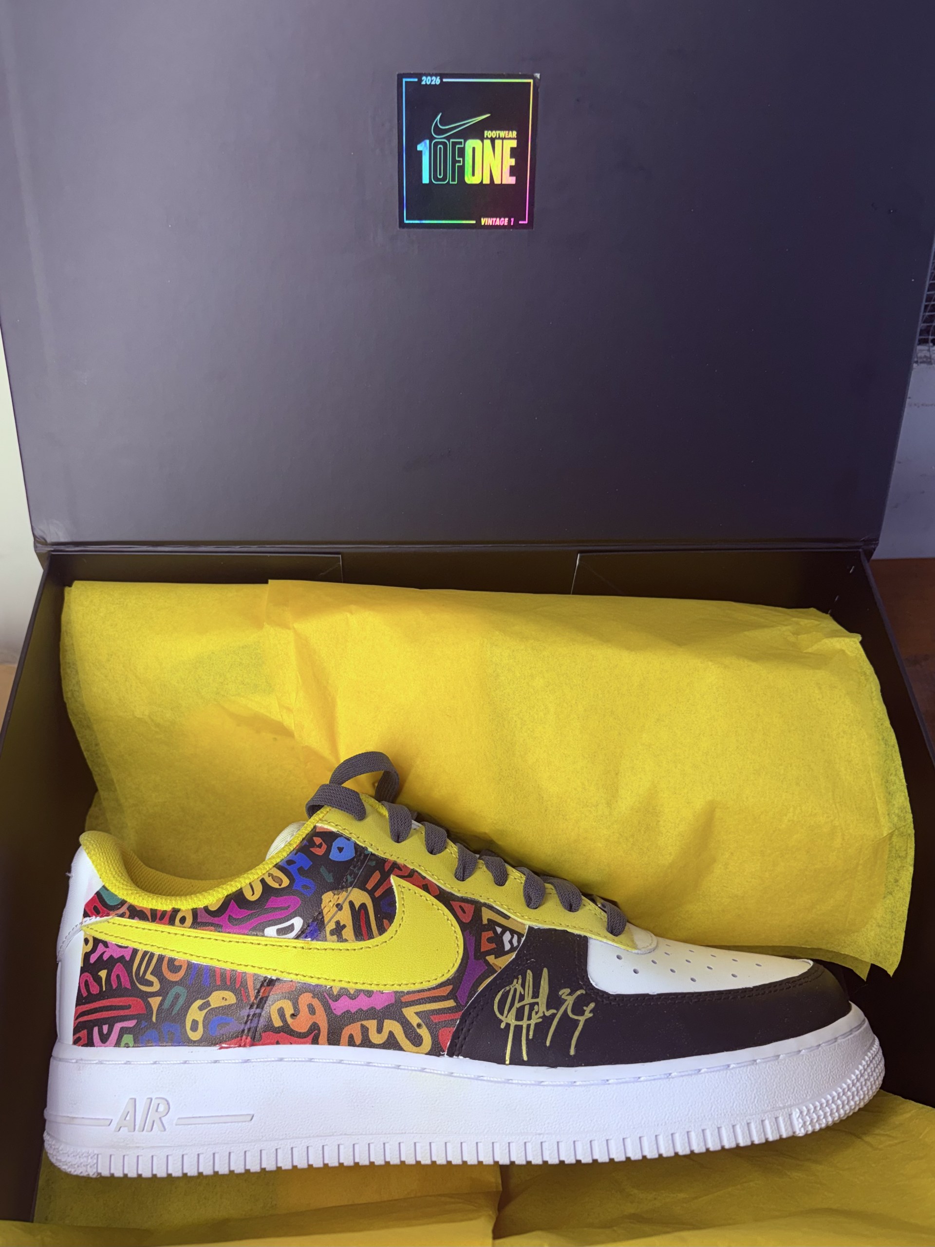 Signed Air Force 1 with yellow swoosh and multicolour doodle print, resting in a 1ofONE presentation box on yellow tissue