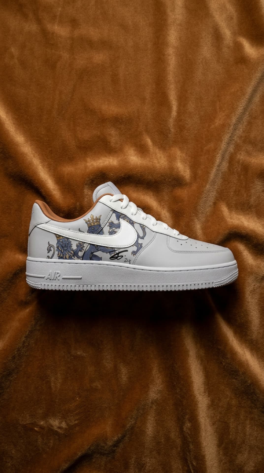 White Air Force 1 with a crowned heraldic lion panel and caramel heel tab, laid on caramel velvet